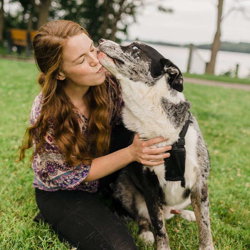 female smiling and hugging australian shepherd dog that's licking her face outdoors by lake